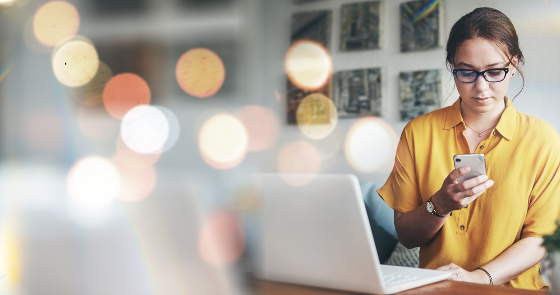 Woman in yellow working at computer with smartphone in hand