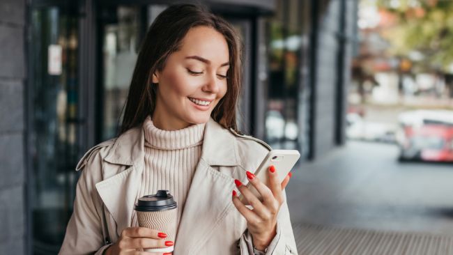 Woman walking with cup of coffee while smiling at smartphone