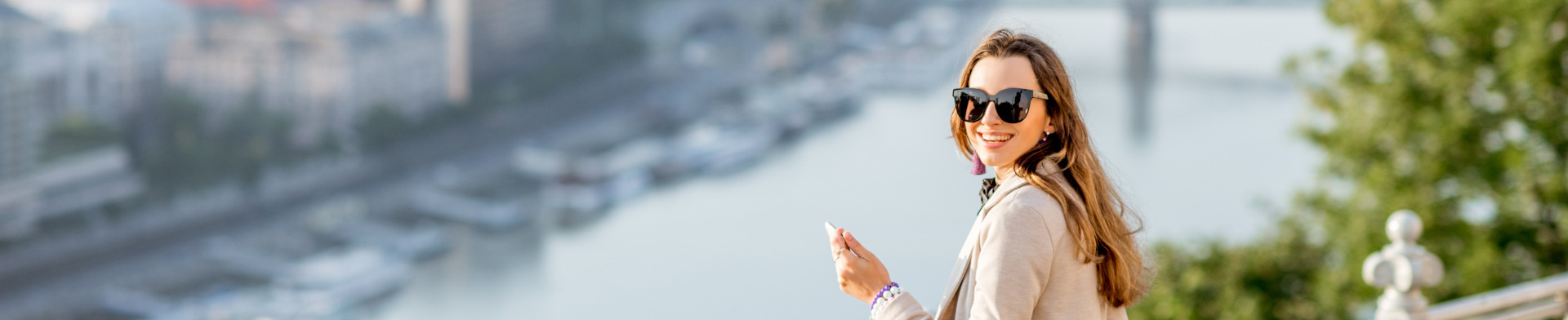 A woman wearing sunglasses with a phone in her hand smiling with a river in the background.